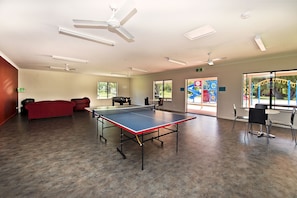 Amplio espacio interior con mesa de ping pong, ventiladores de techo y ventanas grandes que dan vistas a un patio de juegos exterior.