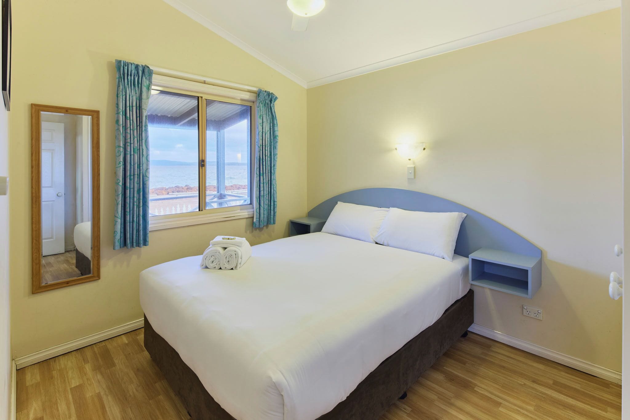 A hotel room with a bed, a nightstand, a wall-mounted lamp, a window with curtains, and a view of the sea.