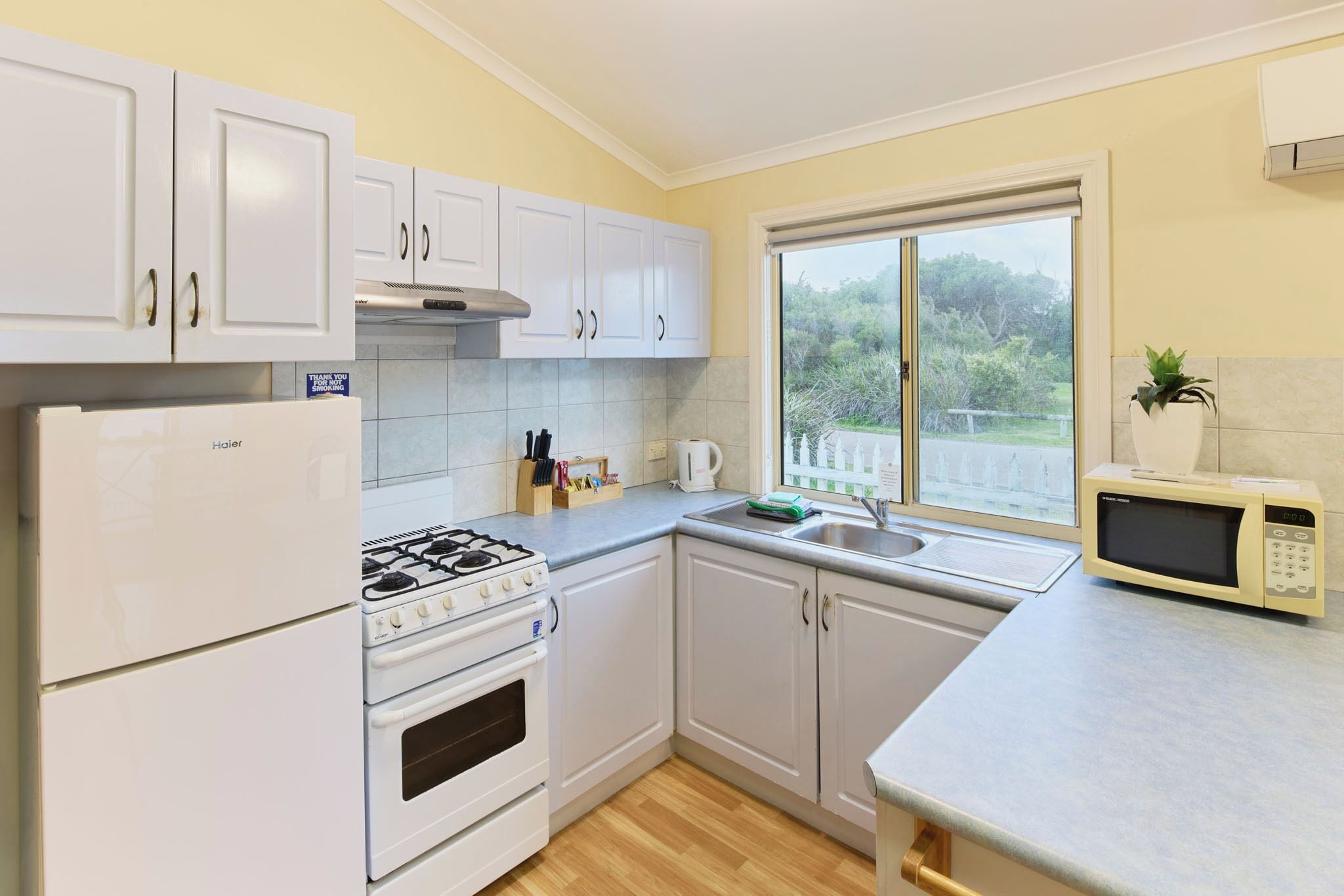 A kitchen with white cabinets, a gas stove, an oven, a microwave, and a window with a view of greenery.
