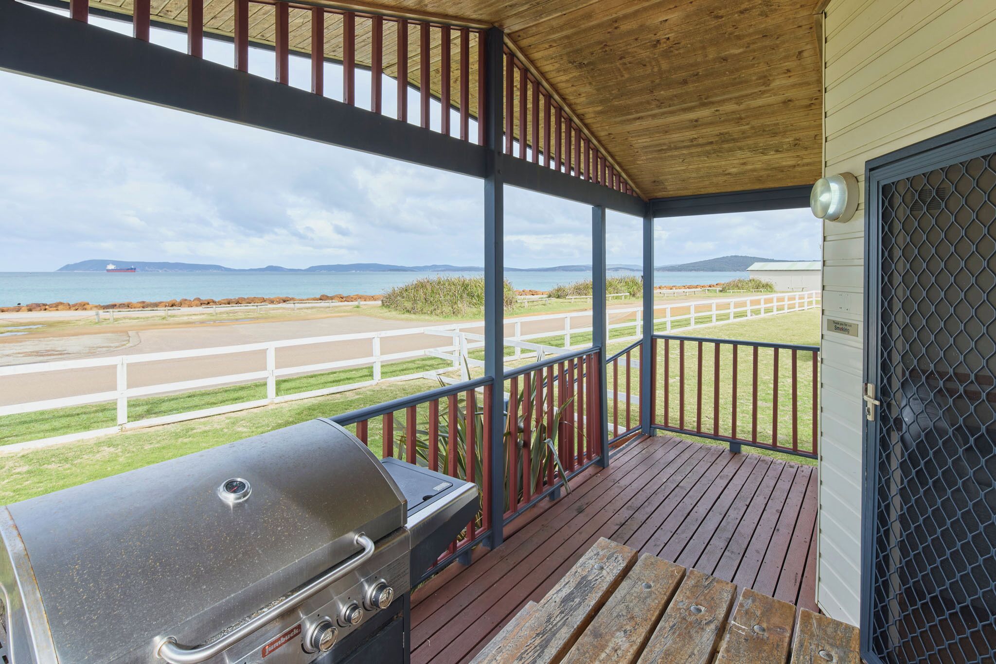 Een overdekt terras met een barbecue, uitzicht op het strand en een witte hek.