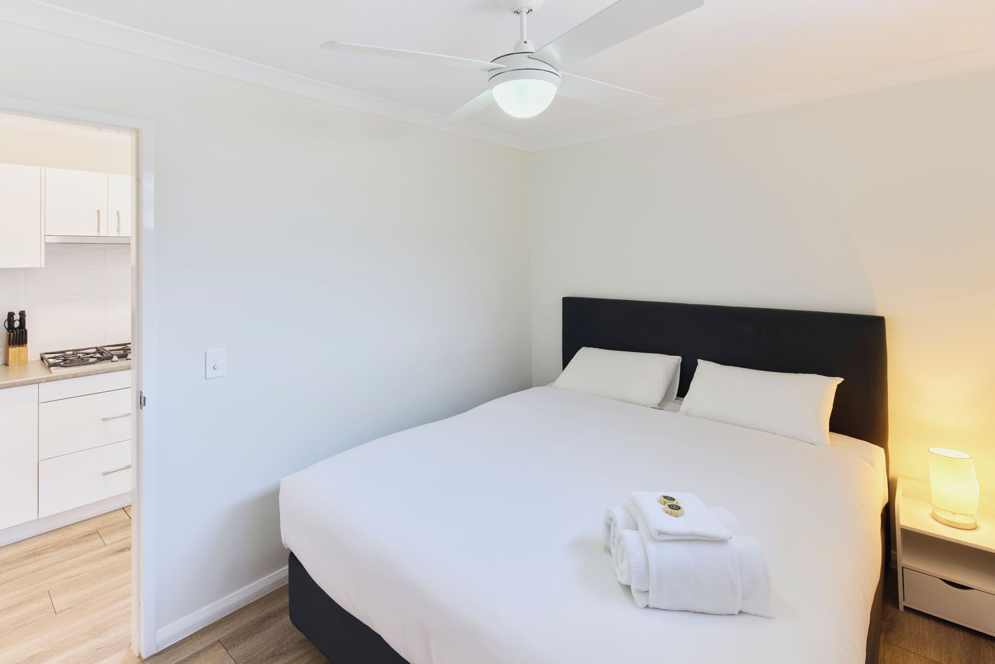 A hotel room with a bed, a nightstand with a lamp, a ceiling fan, and a kitchen area with cabinets and a stove.