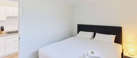 A hotel room with a bed, a nightstand with a lamp, a ceiling fan, and a kitchen area with cabinets and a stove.