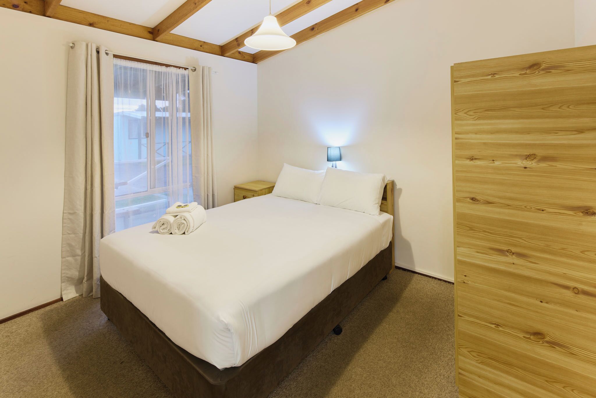 A bedroom with a bed, a nightstand, a lamp, a window with curtains, and a wooden wall.
