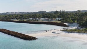 A coastal beach with a rocky breakwater, clear turquoise water, and a few people walking on the sand.