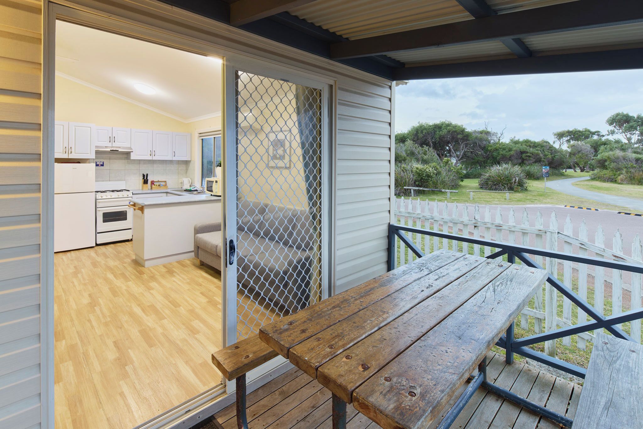 Une terrasse en bois avec une table et des chaises, une porte-fenêtre coulissante donnant sur une cuisine, et une vue sur une route et la verdure.