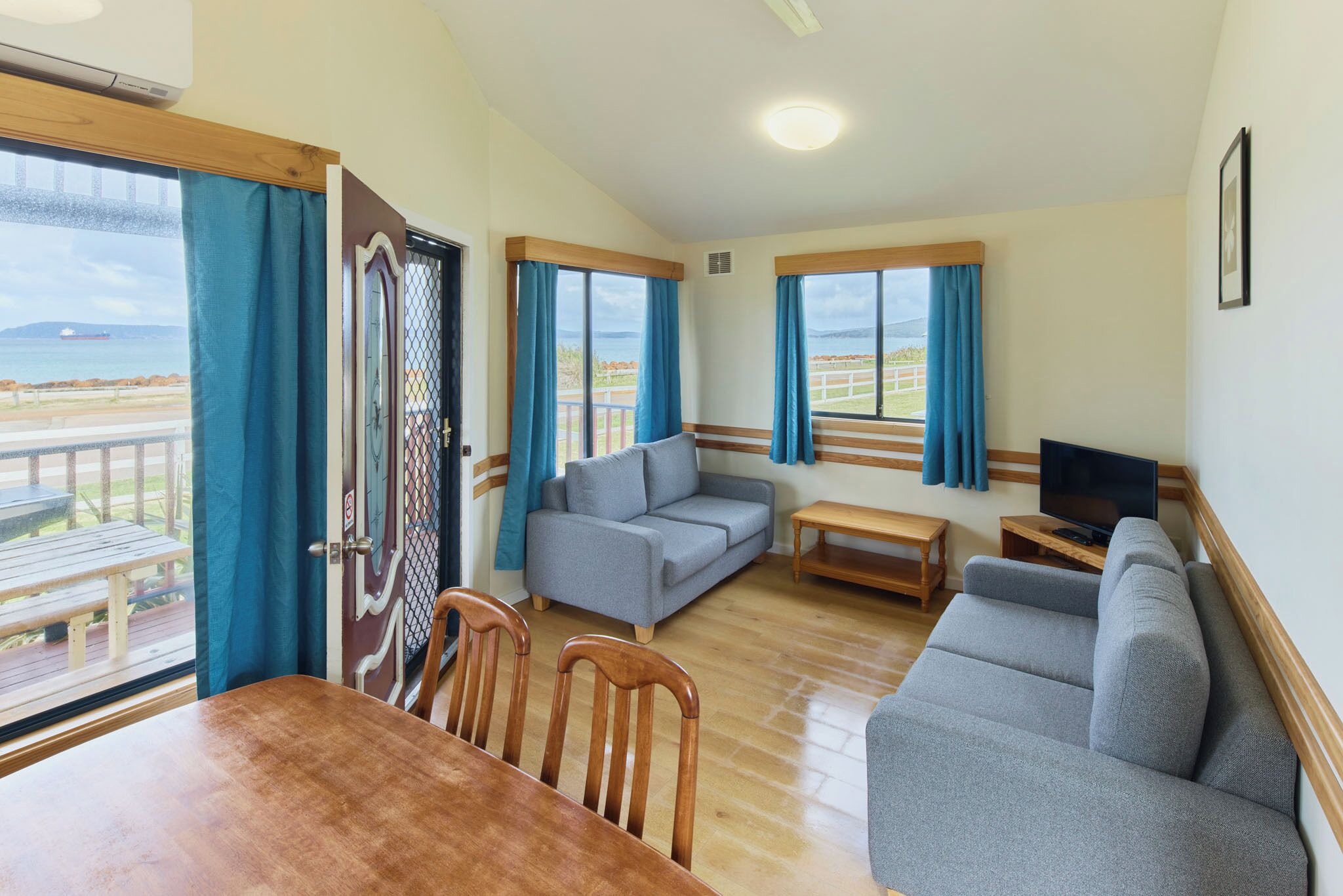 A room with a wooden dining table, two grey sofas, a TV on a wooden stand, and large windows with curtains.