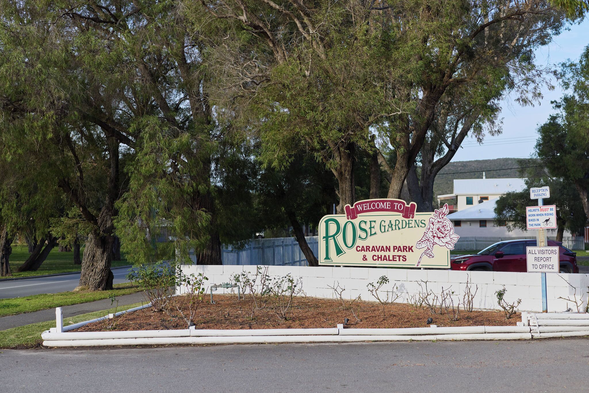 A sign for Rose Gardens Caravan Park & Chalets with a white fence and trees in the background.