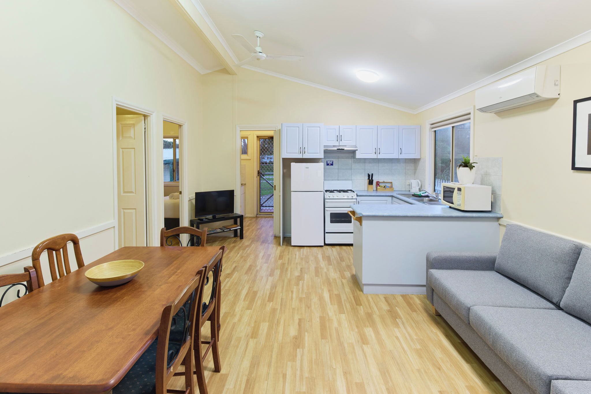 A compact kitchen with white cabinets, a dining table with chairs, a grey sofa, and a TV.