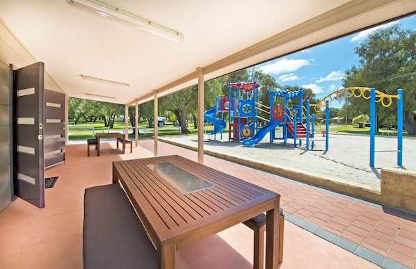 Un espacio de estar al aire libre techado, con mesa y bancos de madera, con vistas a un parque infantil con juegos de colores.