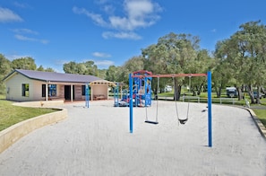 Un parque infantil con columpios y una estructura para escalar, un pequeño edificio con porche, y un estacionamiento con árboles al fondo.
