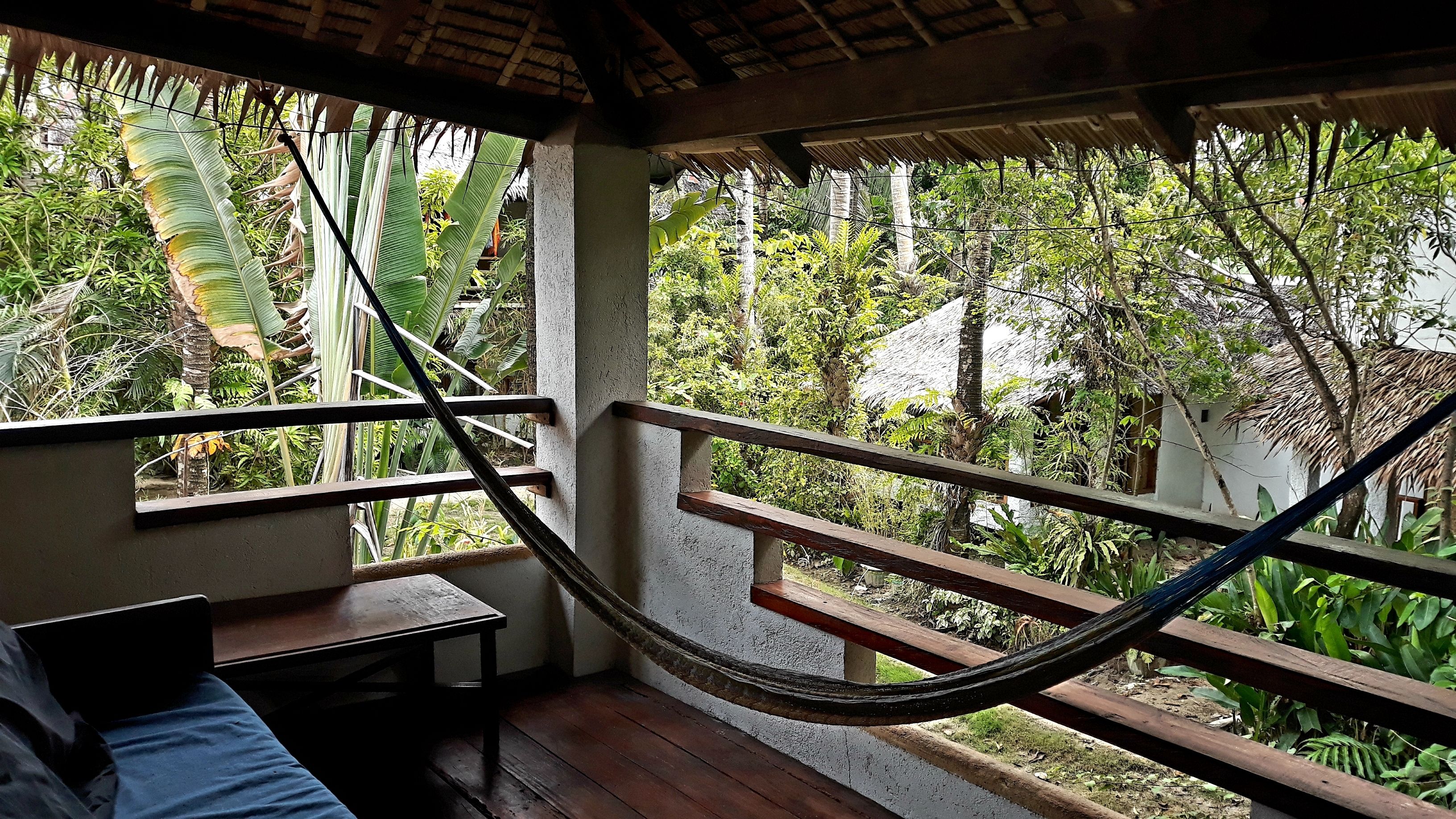 Una hamaca en una habitación con suelo de madera, una cama y una ventana con vista a un jardín exuberante.