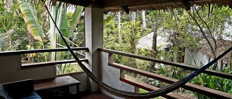 Una hamaca en una habitación con suelo de madera, una cama y una ventana con vista a un jardín exuberante.