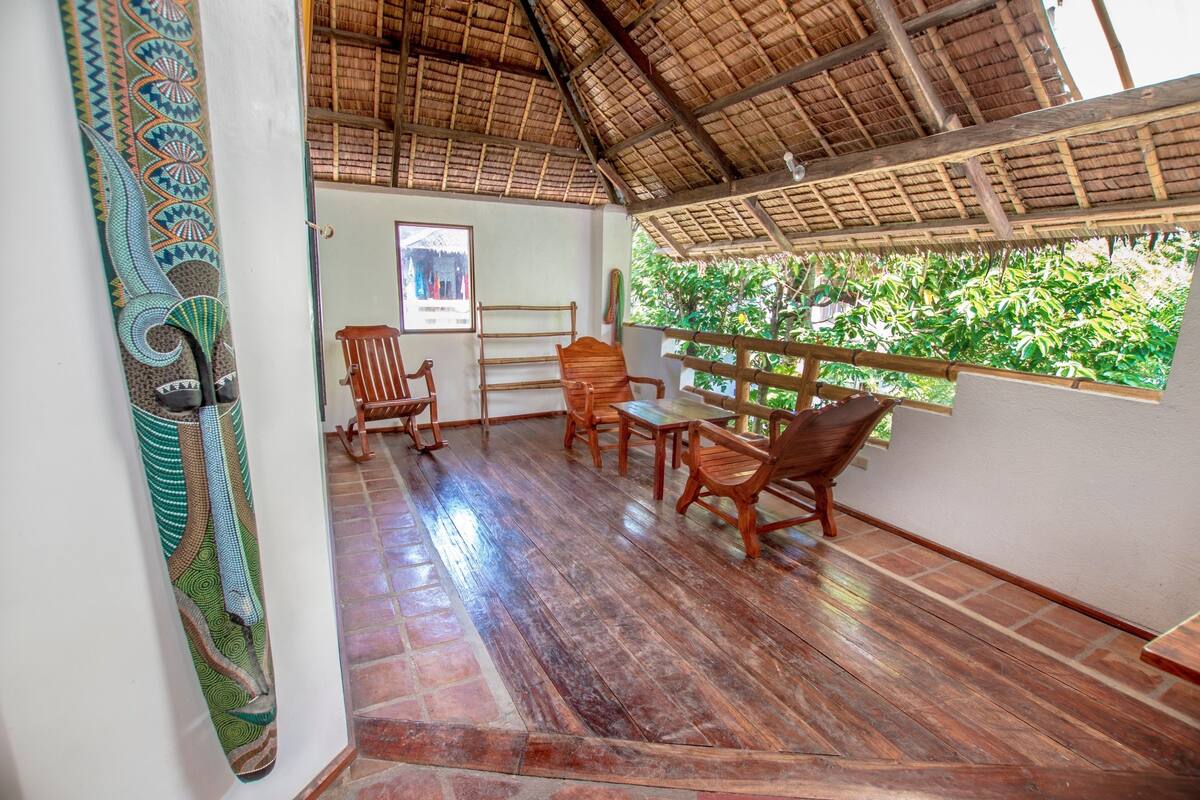 Una habitación con piso de madera, techo de paja y ventana con vista a follaje.