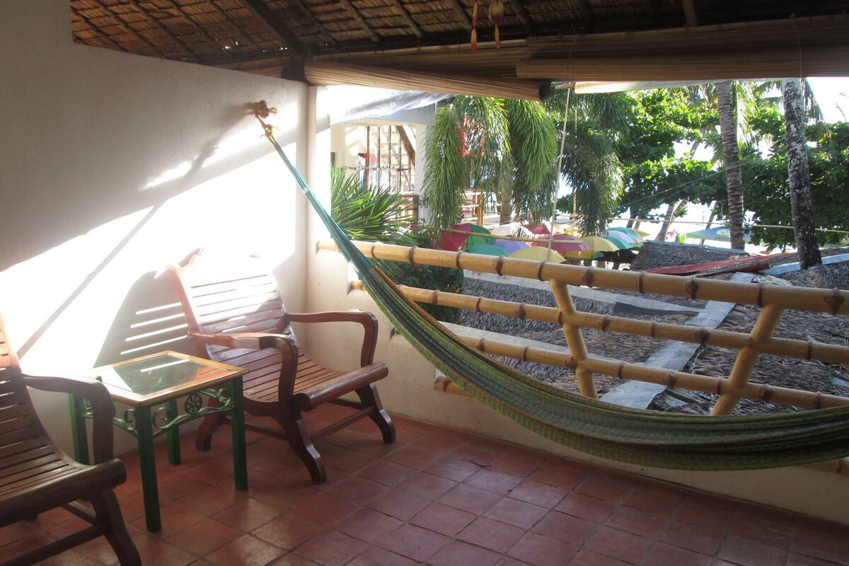 Una silla hamaca y una mesa de madera en un patio con baldosas, con vista a una piscina y sombrillas.