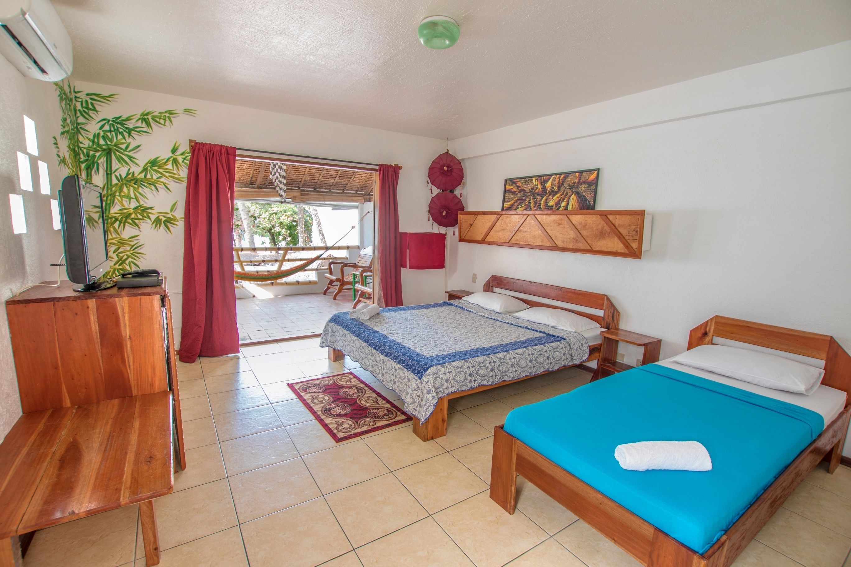 A room with a bed, a wooden bench, a desk, a chair, a potted plant, and a window with curtains.