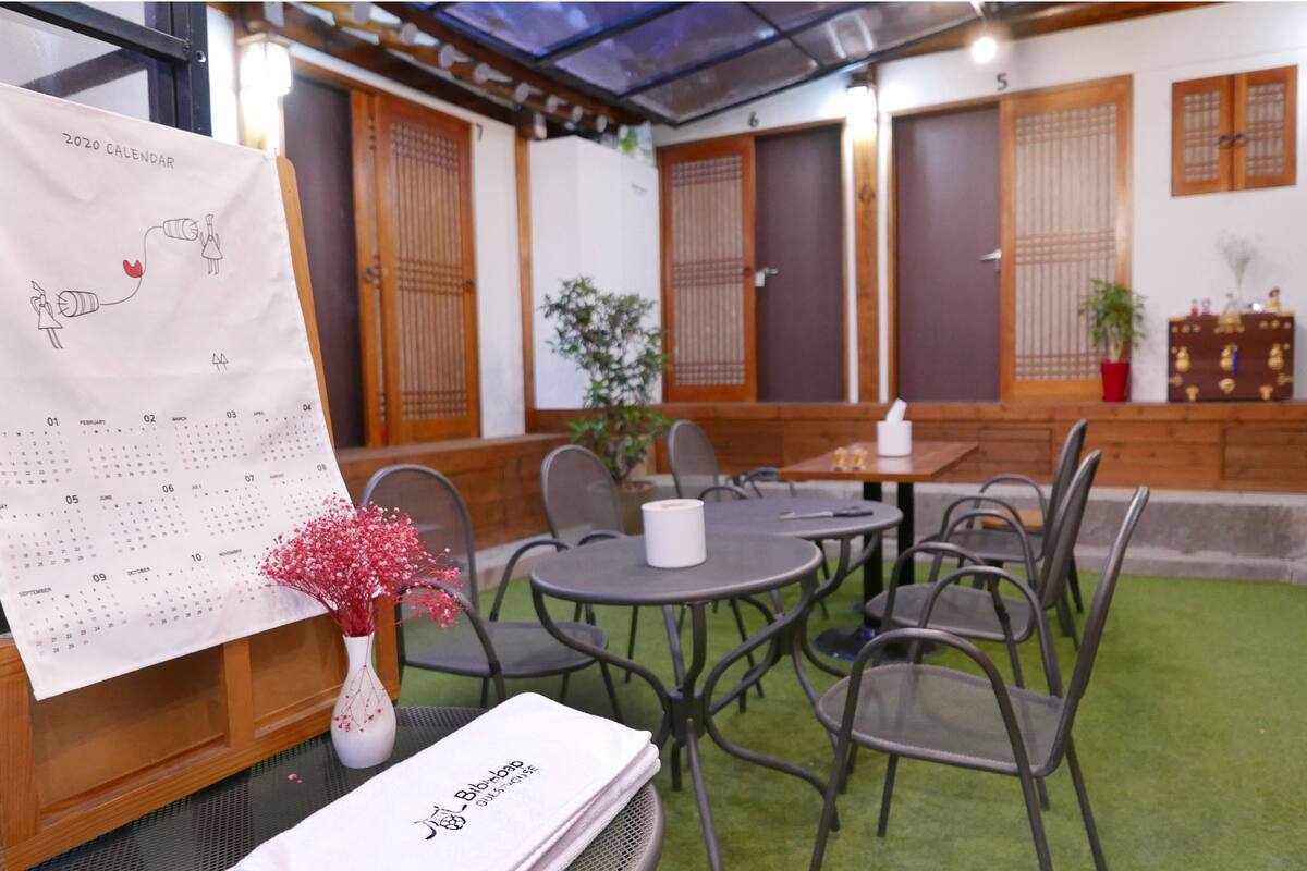 A traditional Japanese-style room with tatami flooring, low tables, and chairs. A calendar is displayed on a stand, and there is a small potted plant on a table.