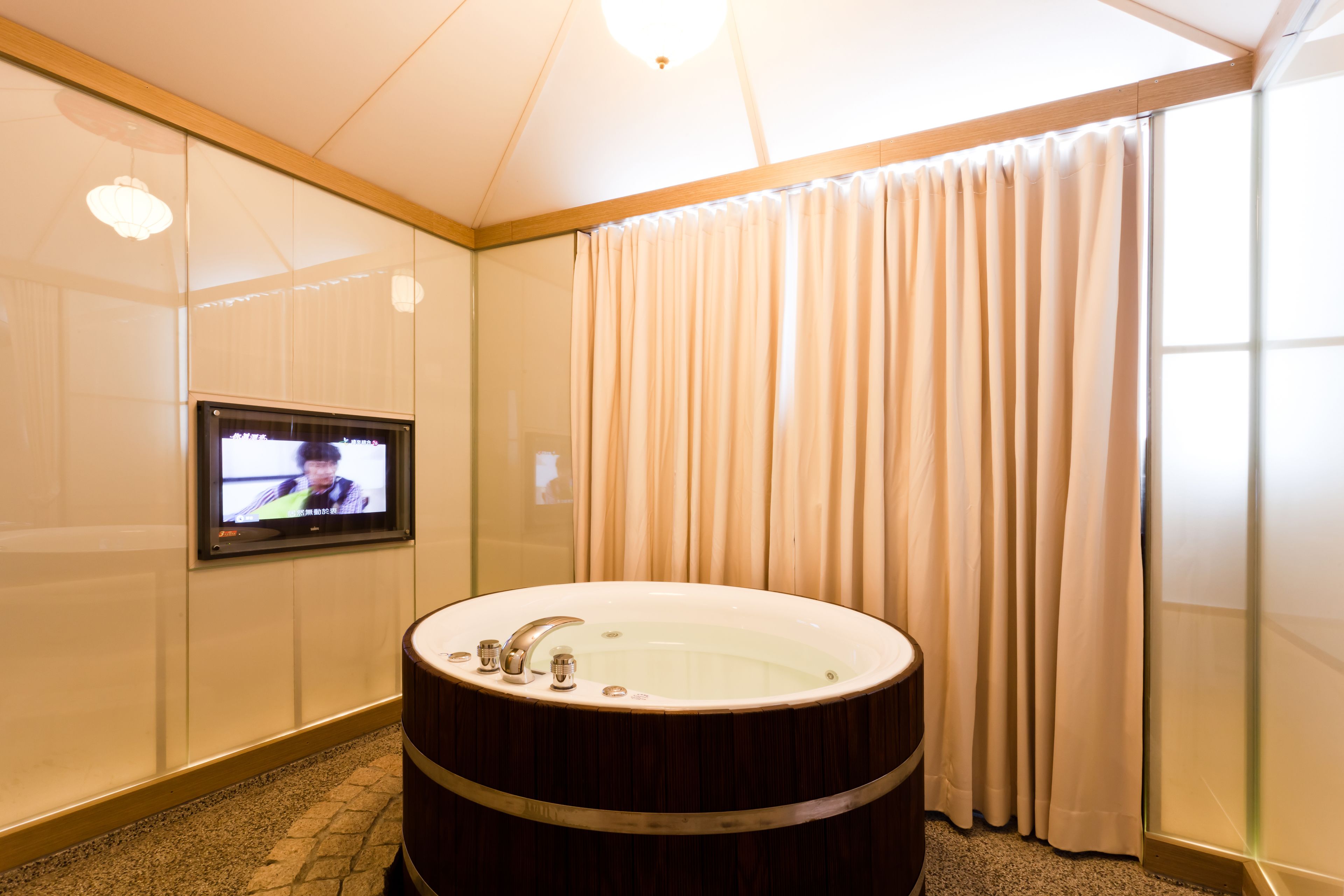 A modern bathroom with a large bathtub, a television mounted on the wall, and a window with curtains.