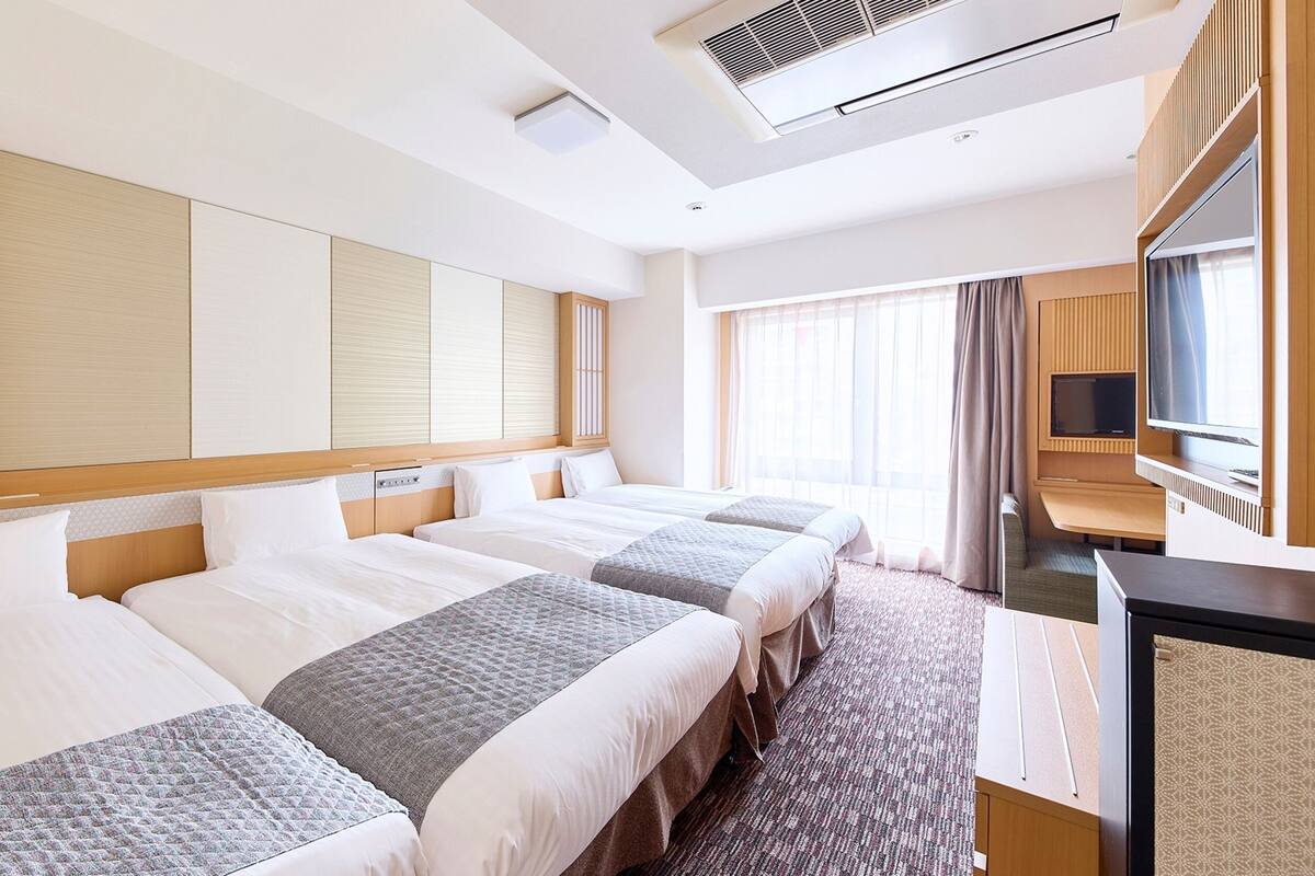A hotel room with three beds, a TV, and a large window with curtains.