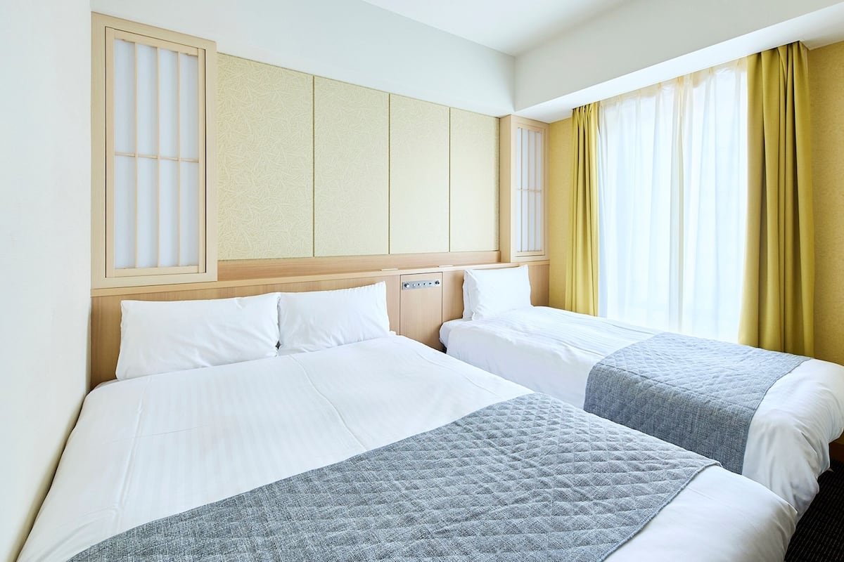 A hotel room with two beds, a wooden headboard, a window with yellow curtains, and a wall with a decorative panel.