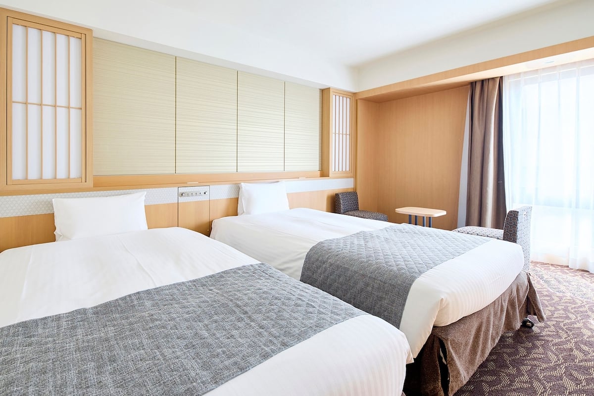 A hotel room with two beds, a small table, a chair, and a large window with curtains.