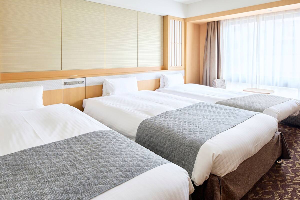 A hotel room with three beds, a wooden headboard, a large window with curtains, and a patterned carpet.