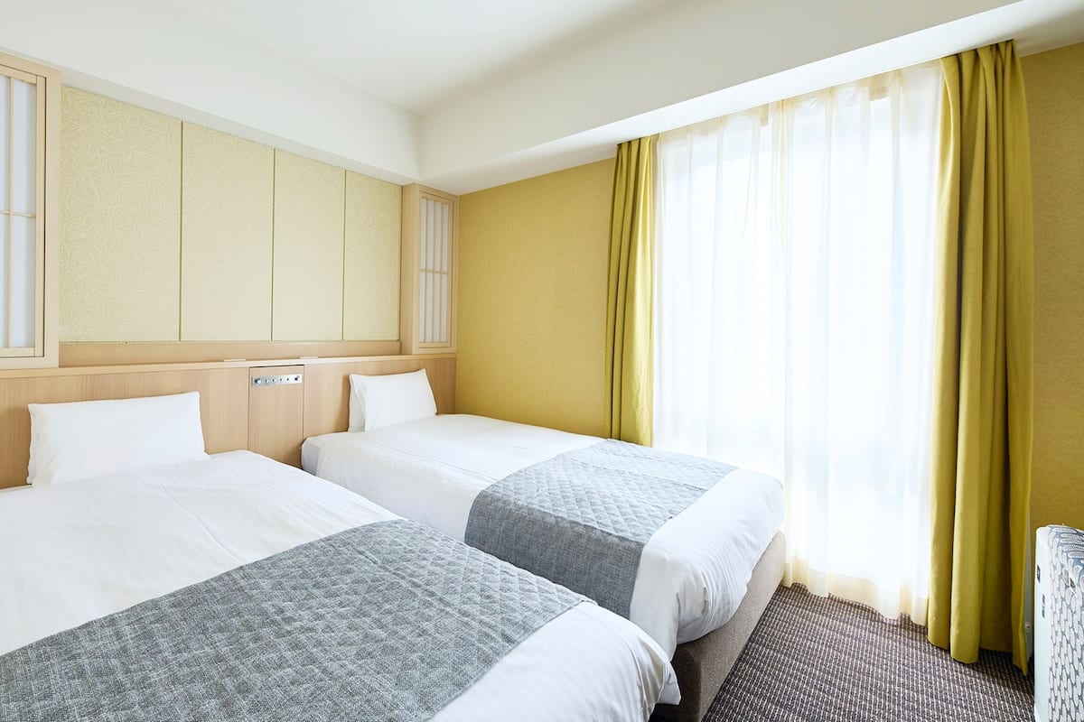 A hotel room with two beds, a wooden headboard, a large window with yellow curtains, and a view of the outside.