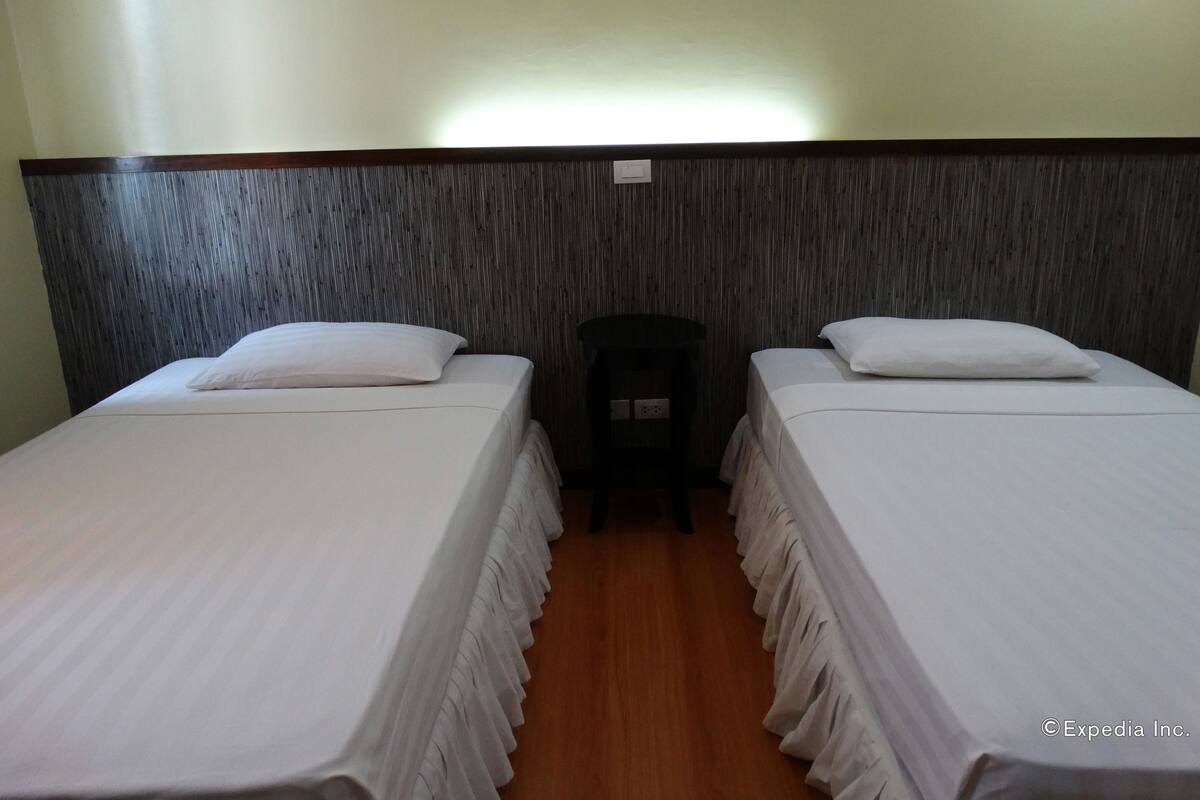 Une chambre d’hôtel avec deux lits simples, une tête de lit en bois, une petite table et une prise de courant au mur.