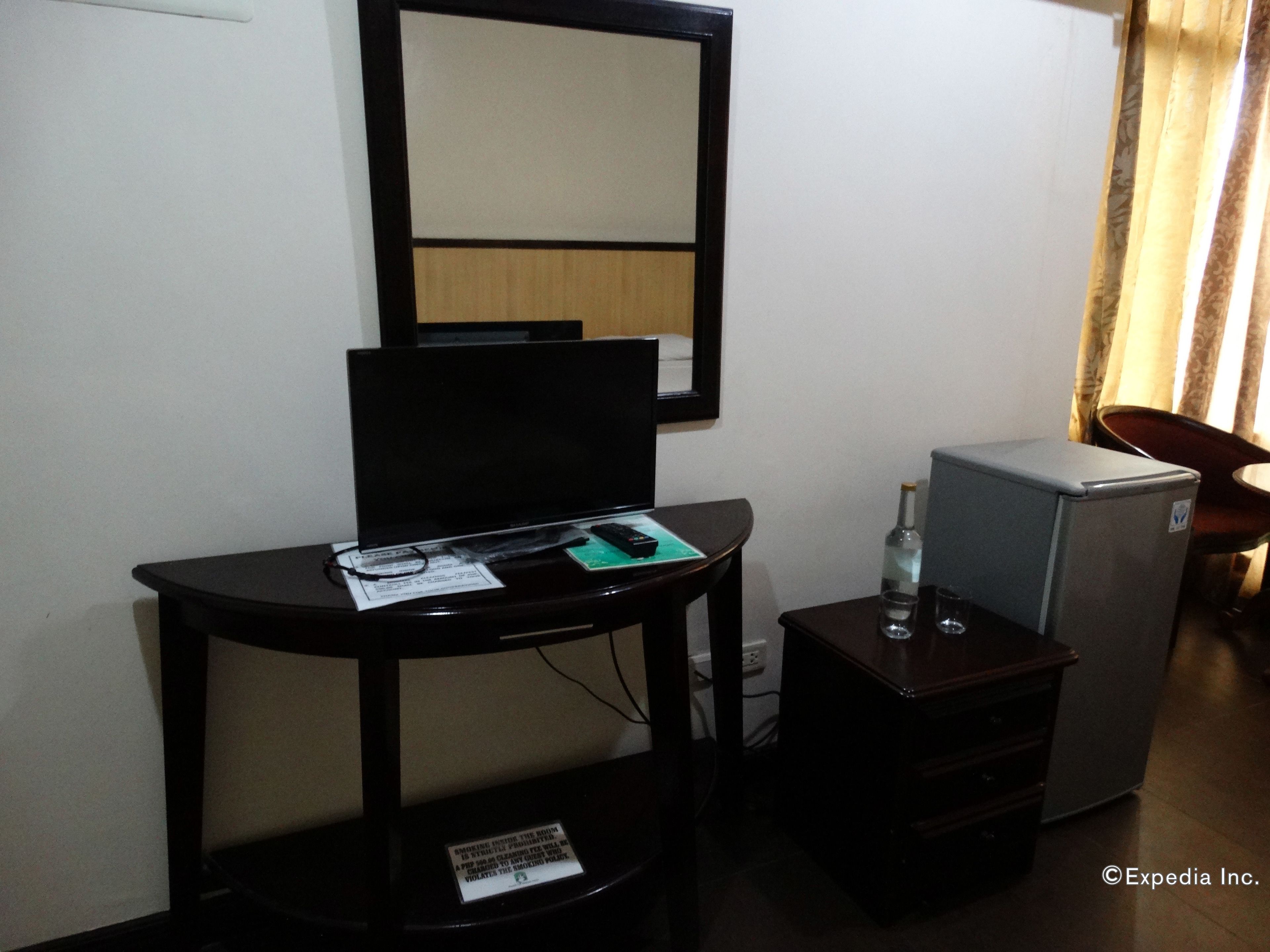 A small hotel room with a desk, a chair, a mirror, a mini-fridge, and a small table with a bottle and glasses.