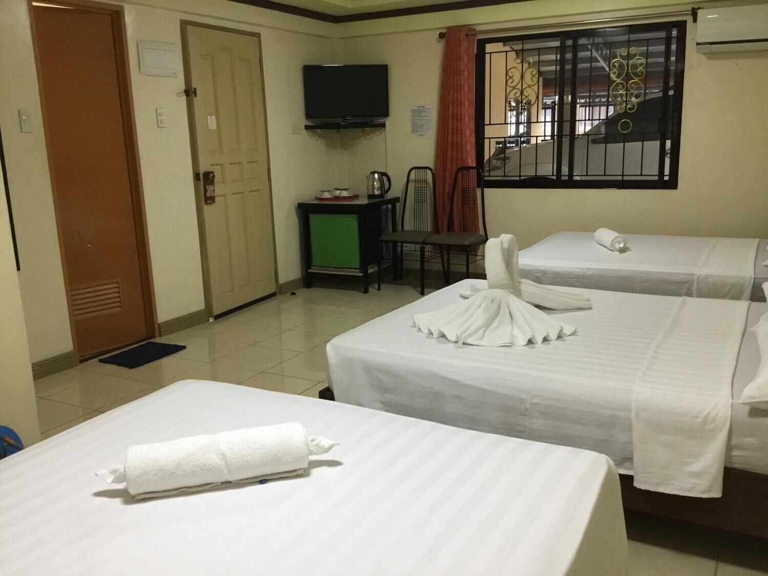 A hotel room with two beds, a television, a small table with a kettle, and a window with bars.