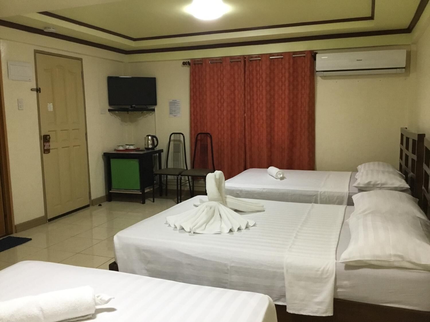 A hotel room with two beds, a TV, a desk with a kettle, and a red curtain.