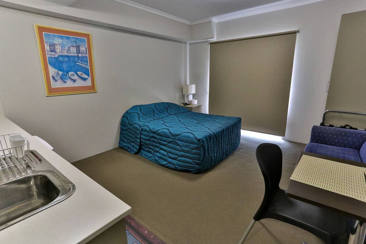 A compact hotel room with a single bed, a small desk, a chair, a sink, and a large window.