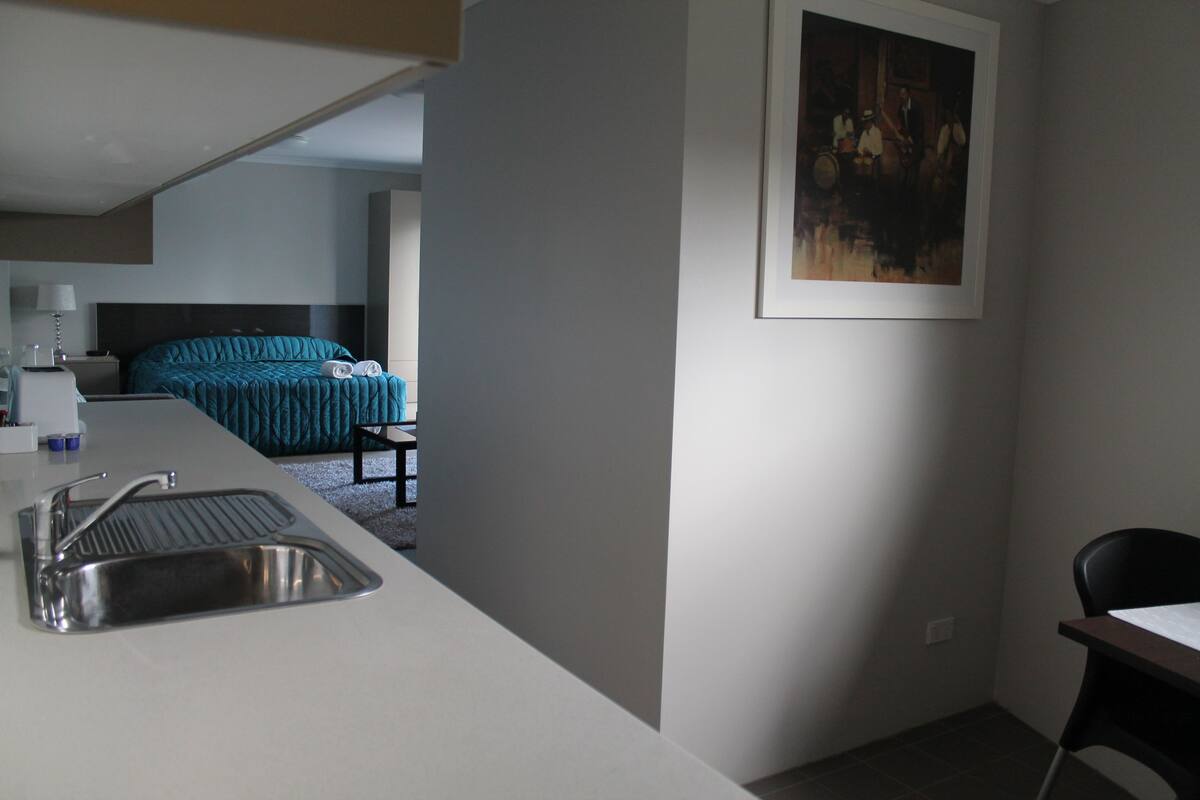 A modern hotel room with a kitchenette, a bed with teal bedding, and a framed painting on the wall.