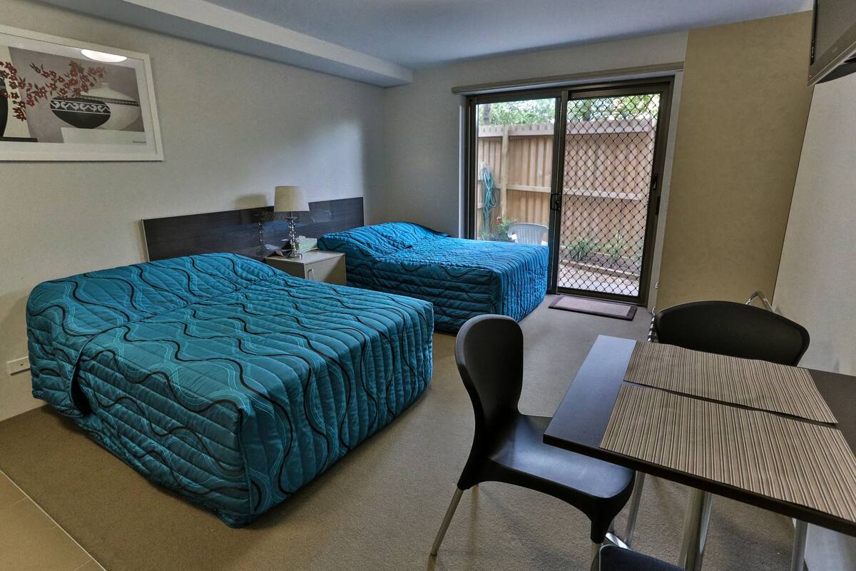 A hotel room with two beds, a table, a chair, and a sliding glass door to the outside.