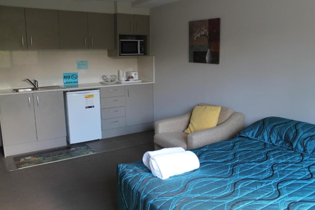A compact room with a kitchenette, a bed with a blue patterned bedspread, a chair with a yellow pillow, and a small refrigerator.