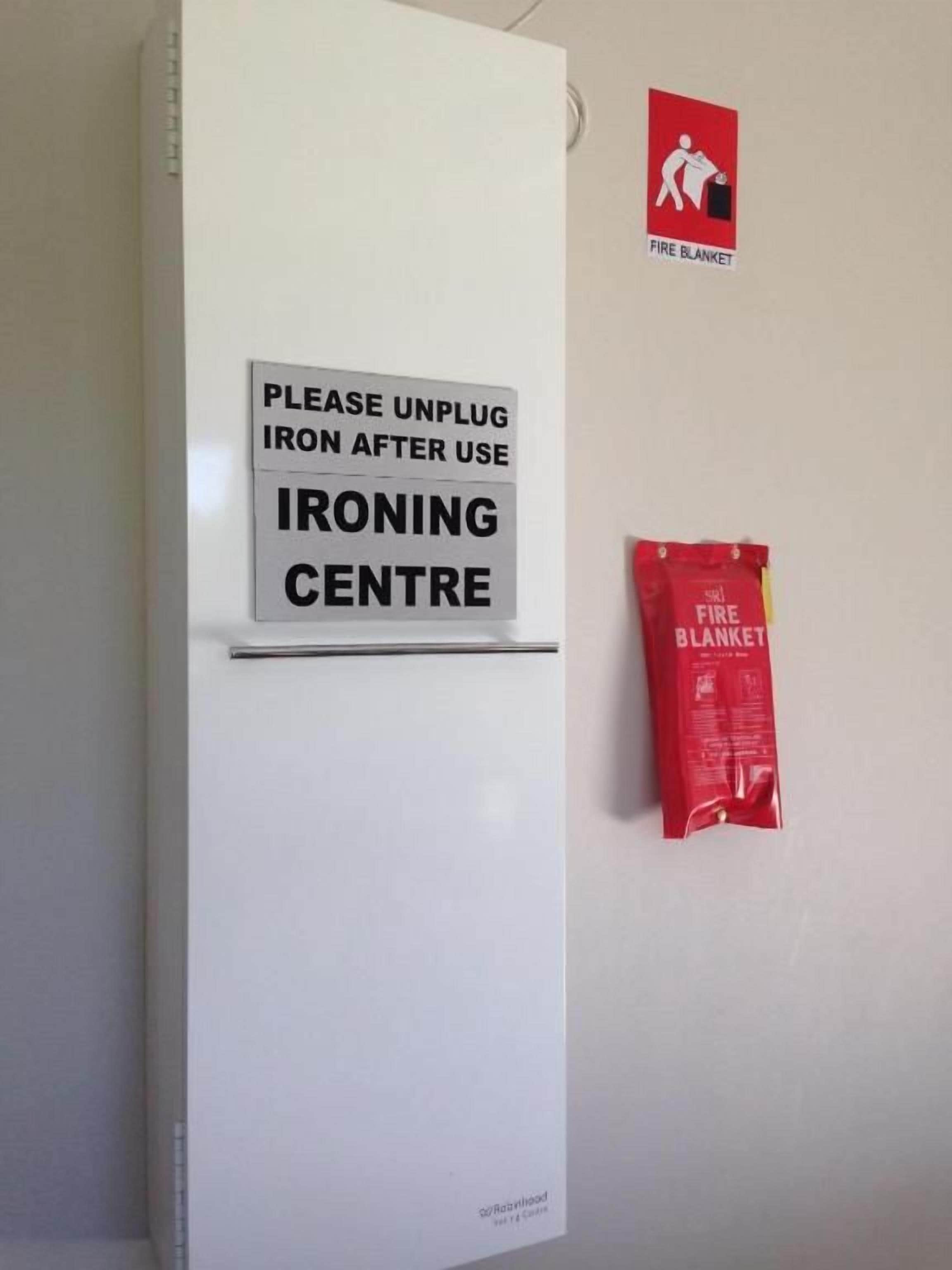 A white wall with a sign that reads 'Please unplug iron after use' and a fire blanket hanging on the wall.