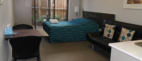 A compact hotel room with a bed, a small desk, a chair, a sofa, and a kitchenette.