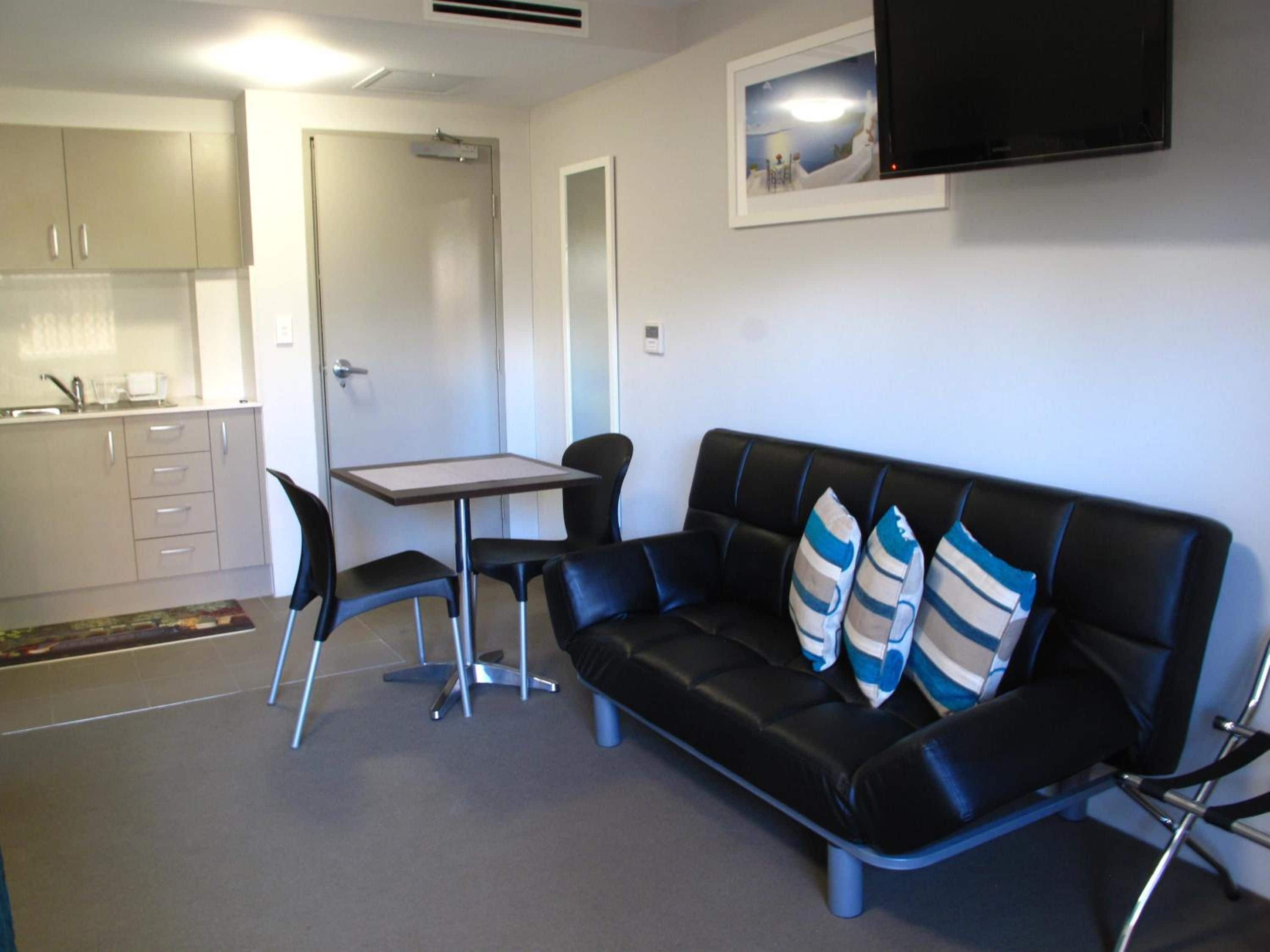 Un espace de vie compact comprenant un coin cuisine, un canapé en cuir noir avec des coussins à rayures, une petite table et un téléviseur à écran plat fixé au mur.
