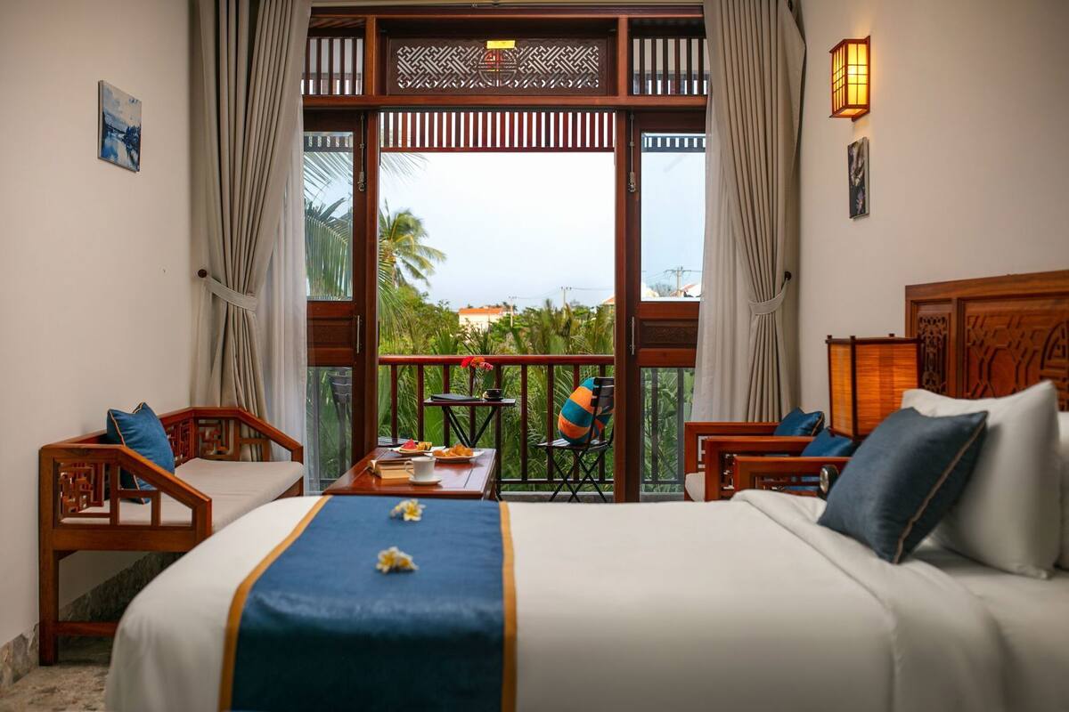 Ein Hotelzimmer mit Bett, zwei Stühlen, einem kleinen Tisch mit einer Speisewagenplatte und einem Balkon mit Blick auf Grünflächen und Gebäude.