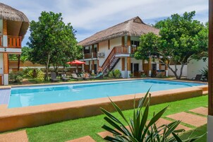 Una piscina rodeada por un jardín exuberante, con un edificio que tiene techo de paja y una escalera que conduce a un balcón.