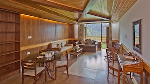 A spacious living area with wooden ceilings, a sofa, a dining table with chairs, and a large window offering a view of the outdoors. - Vivaan The Sunrise Resort (Manali)