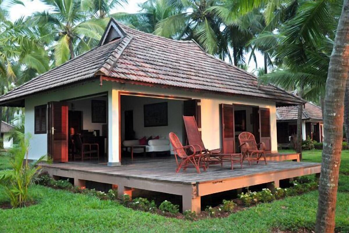 Uma casa térrea com telhado de telhas, varanda com cadeiras e um pequeno jardim.