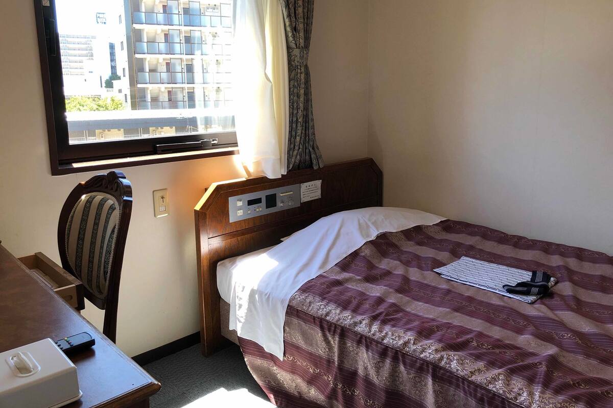 A hotel room with a single bed, a wooden headboard, a desk with a chair, a window with curtains, and a view of buildings outside.