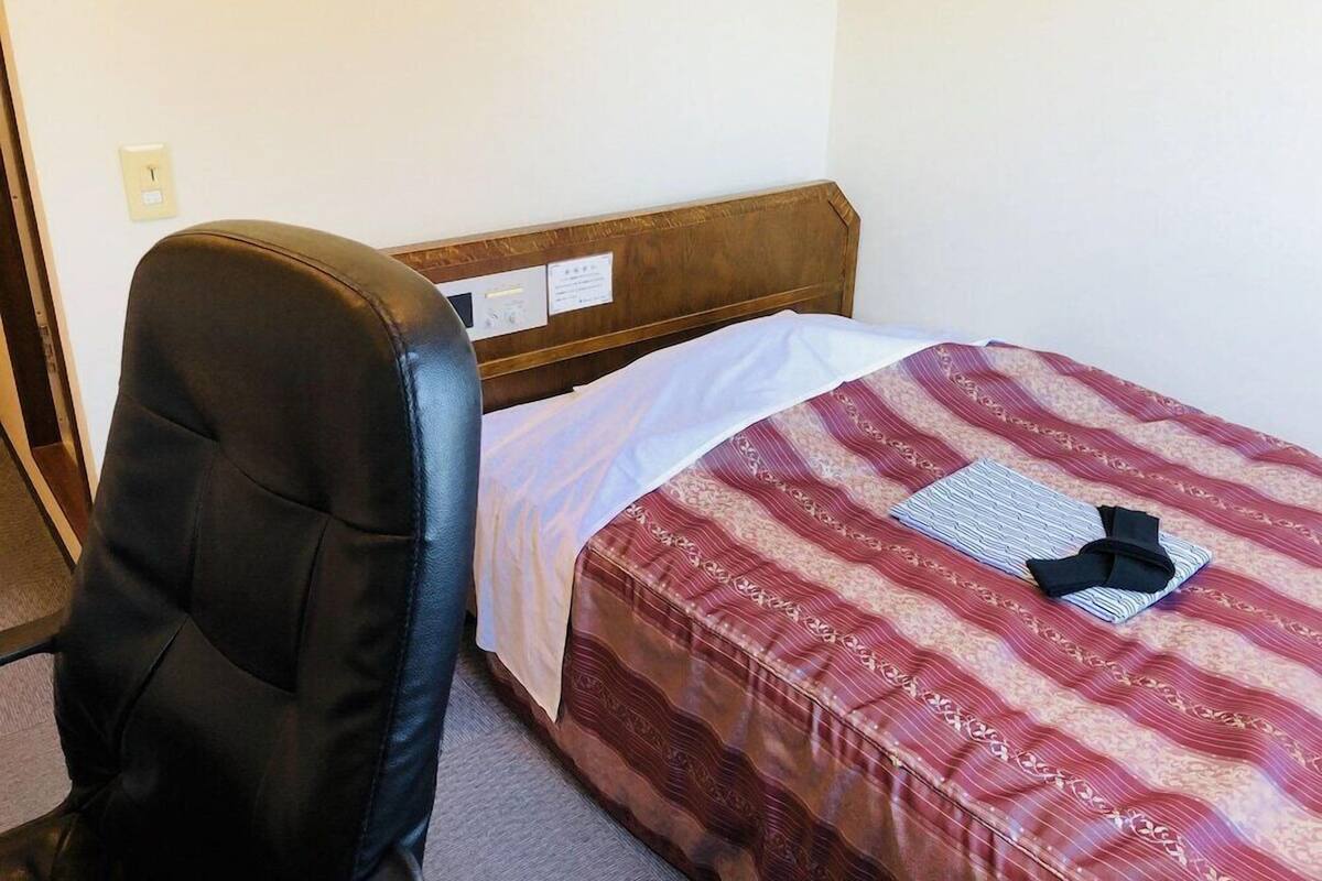 A hotel room with a single bed, a wooden headboard, a black leather chair, and a white wall with a light switch.