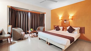 A hotel room with a bed, two chairs, a small table with a fruit bowl, a ceiling fan, and a window with curtains. - Hotel Orchard Pune (Pune)