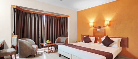 Habitación de hotel con cama, dos sillas, una mesita con frutero, ventilador de techo y ventana con cortinas.