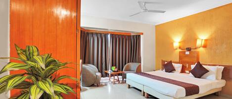 Quarto de hotel com cama, escrivaninha, duas cadeiras, planta em vaso, ventoinha de teto e janela com cortinas.
