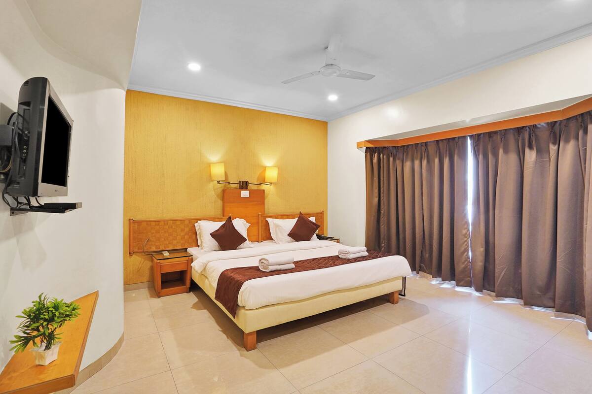 A hotel room with a large bed, a television mounted on the wall, a ceiling fan, and a window with curtains.