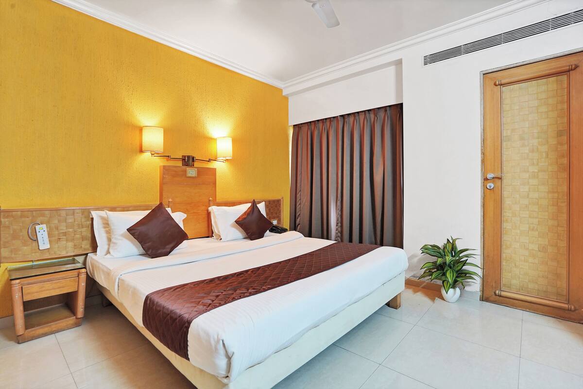 A hotel room with a large bed, two bedside lamps, a wooden headboard, a nightstand, a potted plant, and a door with a textured panel.