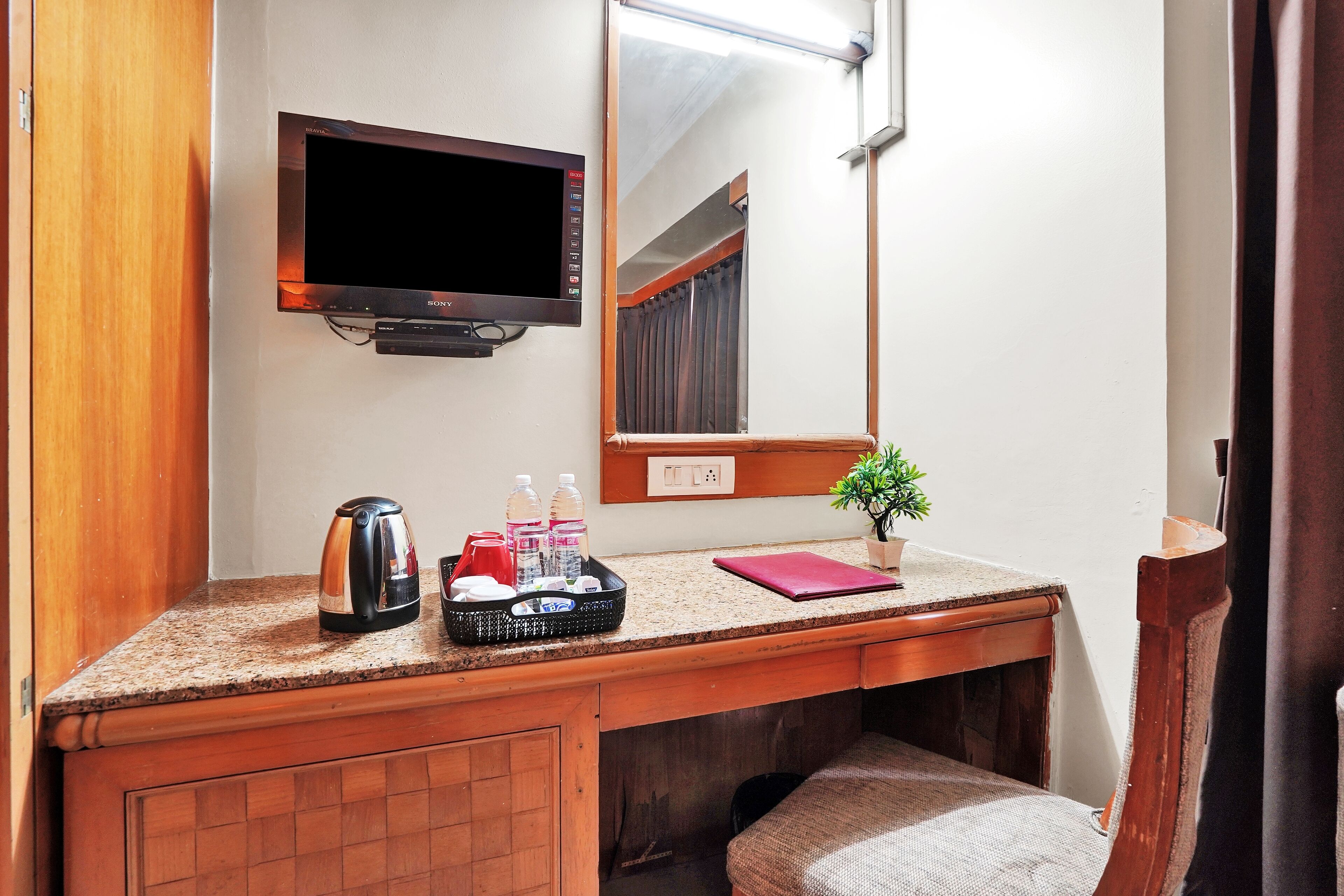A small bathroom with a wooden vanity, a sink, a mirror, a television mounted above the mirror, and a small potted plant on the vanity.