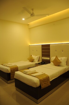 A hotel room with two beds, each with a headboard, a bedside table, and a ceiling fan. - Saibala Grand Hotel (Chennai)